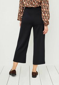 Pantaloni neri a gamba larga con una finitura testurizzata e vita elasticizzata, abbinati a scarpe in camoscio marroni, visti da dietro.