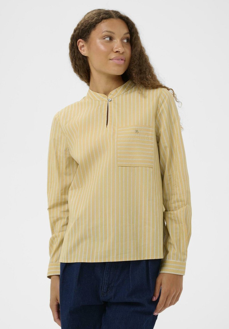 Jeune femme portant une blouse rayée jaune à manches longues avec une poche poitrine, se tenant devant un fond blanc uni.