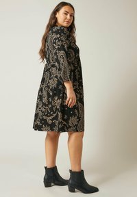 Schwarzes Kleid mit beigem Paisleymuster, mit Dreiviertelärmeln und tailliertem Bund, getragen mit schwarzen Knöchelstiefeln.