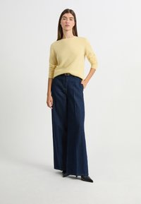 Żółty, dziany sweter z długimi rękawami, w połączeniu z wysokim stanem, szerokimi ciemnoniebieskimi dżinsami. Czarne szpilki z ostrym noskiem dopełniają stylizację.