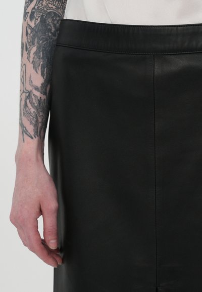 Fustă din piele neagră cu textură netedă și un design drept. Are un tiv simplu și cusături laterale. O mână odihnindu-se pe material.