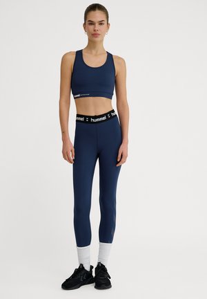 Marineblauwe sport-bh en leggings met een zwarte tailleband waarop een logo staat. De leggings hebben een subtiel patroon aan de zijkant en zitten nauwsluitend.