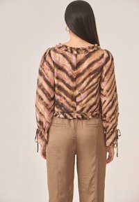 Top largo de manga larga traslúcido marrón con rayas onduladas rosas y marrones, cuello con volantes y acentos de mangas atadas, combinado con pantalones marrones claros.