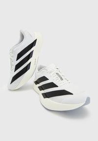 Paire de chaussures de course blanches avec des rayures noires, des semelles épaisses et rembourrées, et "ADIZERO" écrit sur la languette, présentée sur un fond uni.