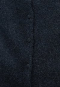 Detailní záběr na tmavou načechranou látku se čtyřmi zakrytými knoflíky seřazenými vertikálně uprostřed.