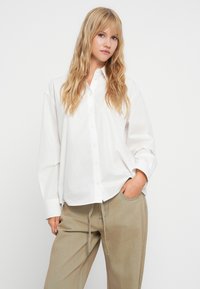 Λευκή υπερμεγέθη πουκάμισο με κουμπιά και μακριά μανίκια, φτιαγμένο από ελαφρύ ύψιστο. Συνδυασμένο με μ beige παντελόνια με κορδόνι και τσέπες.