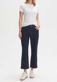 Jeans a zampa d'elefante in denim blu scuro con orlo grezzo, tasche frontali e cuciture a contrasto, abbinati a una maglietta bianca a righe aderente e scarpe da ginnastica.