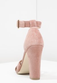 Rosa mocka högklackad sandal med ankelsnörning och guldfärgad spänne, blockklack och öppen tå, visad bakifrån på en vit yta.