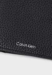 Juodas tekstūruotas odinis piniginė su matomu sidabriniu "Calvin Klein" logotipu. Yra siuvinėta kraštinė ir kompaktiškas stačiakampio formos dizainas.