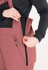 Weinrote Latzhose aus strukturiertem Material, mit hohem Kragen und Reißverschlussöffnungen. Mit schwarzen Akzenten und einer seitlichen Tasche mit Reißverschluss.