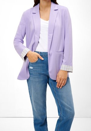 Vrouw draagt een lavendelkleurige blazer met opgerolde gestreepte manchetten over een wit overhemd en blauwe spijkerbroek, één hand in de zak, staand tegen een witte achtergrond.