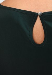 Tissu vert foncé à la texture lisse, avec une petite découpe en forme de trou de serrure à l'arrière, fermée par un bouton argenté.