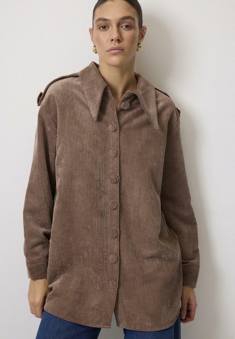 Femeie purtând o cămașă maro din catifea cu nasturi, cu guler larg și cercei aurii, stând pe un fundal simplu și deschis la culoare.