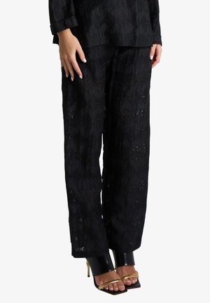 Pantaloni neri a gamba larga con texture indossati con sandali con tacco alto neri e dorati, mostrando piedi e mani con smalto chiaro.
