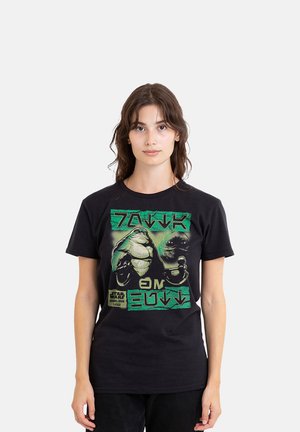 Young woman with brown hair wears a black Star Wars T-shirt featuring Jabba the Hutt and green alien text on a plain background.