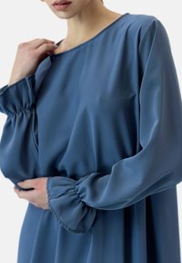 Niebieska sukienka z długimi rękawami, uszyta z gładkiego materiału, z szerokim dekoltem i marszczonymi mankietami, w luźnym kroju i minimalną ilością detali.