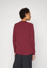 Chemise à manches longues en tissu bordeaux, avec un col rond, des manches raglan et un ourlet légèrement courbé à l'arrière.