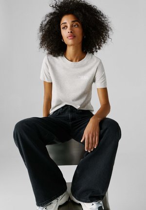 Γυναίκα με σγουρά μαλλιά κάθεται σε καρέκλα, φορώντας ανοιχτό γκρι μπλουζάκι, μαύρο τζιν με φαρδιά πόδια και λευκά αθλητικά παπούτσια.