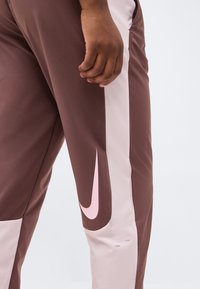 Καφέ και ανοιχτό ροζ αθλητικό παντελόνι με μεγάλο ροζ σήμα Nike στο μηρό και το χέρι ενός ατόμου να ακουμπάει δίπλα από το πόδι.