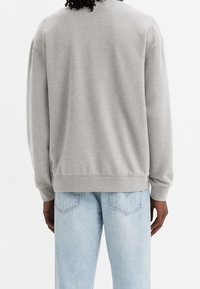 Sudadera gris de manga larga, con cuello redondo y puños acanalados, combinada con jeans de color azul claro que presentan detalles de costura en los bolsillos traseros.