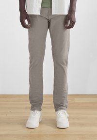 Chinos beiges en coton avec une coupe droite. Portés avec un t-shirt vert et des baskets blanches, sur un fond de sol en bois.