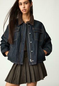 Denimtakki ruskealla corduroy-kauluksella, vetoketjukiinnitys ja kaksi etutaskua. Päälle on puettu tummasta kankaasta valmistettu, pystyraitainen hame.