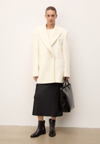 Chaqueta de lana blanca de corte oversize, falda midi de satén negra, botas de cuero negras hasta el tobillo con cremalleras y un bolso tote negro brillante. Diseño minimalista.