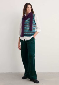 Gilet di maglia con motivo geometrico multicolore, abbinato a una camicia bianca e pantaloni in velluto a coste verde scuro. Gli accessori includono una sciarpa viola.