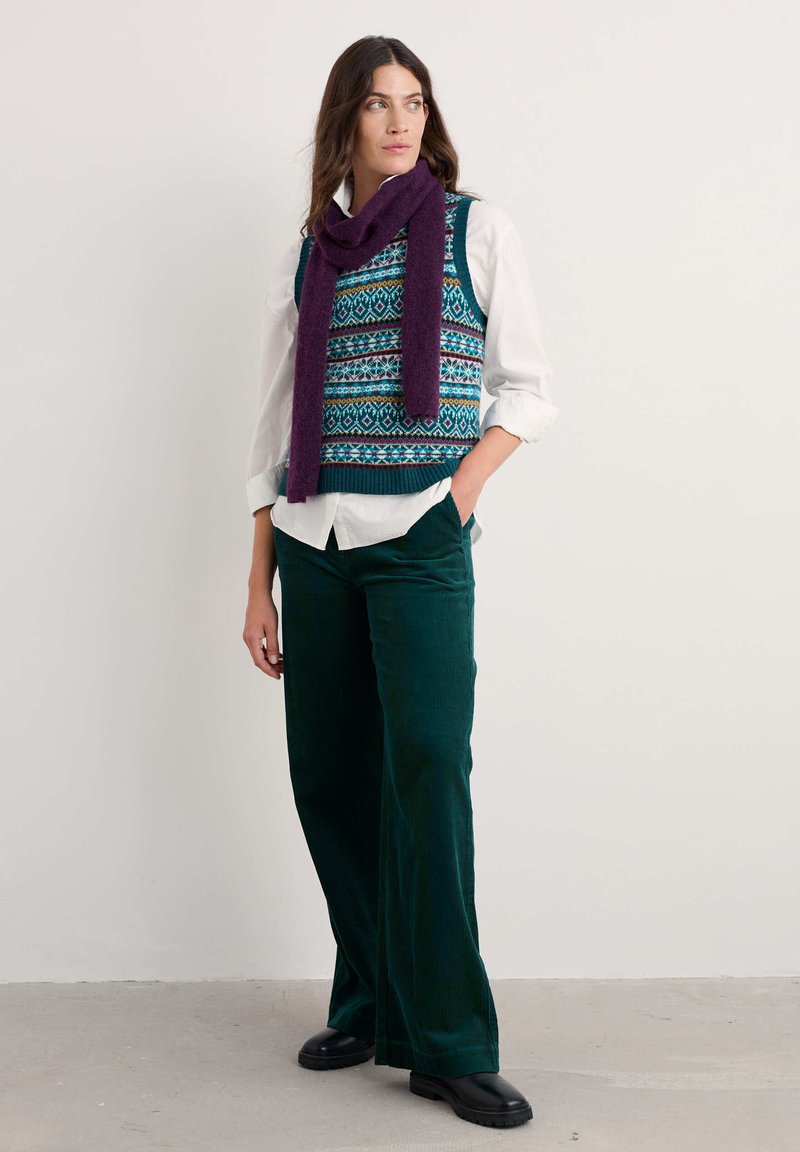 Gilet di maglia con motivo geometrico multicolore, abbinato a una camicia bianca e pantaloni in velluto a coste verde scuro. Gli accessori includono una sciarpa viola.