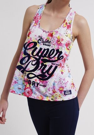 Woman wearing a sleeveless, floral print Superdry tank top with large navy logo text, paired with dark blue fitted pants.