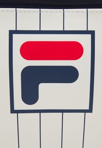 Bolsa de marca FILA con fondo blanco y finas rayas en color navy, un destacado logo en rojo y navy con un diseño geométrico, y cierre de cremallera.