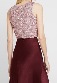 Nainen, jolla on päällästä sleeveless-mekko, jossa on vaaleanpunaisista paljeteista tehty yläosa ja sileä, tumma burgundinpunainen hame, näkyy takaa kädet rentona sivuilla.