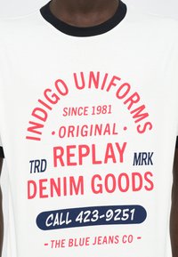 Λευκό μπλουζάκι με κόκκινο και ναυτικό κείμενο. Μαύρο ριπ λαιμόκοψη. Το κείμενο περιλαμβάνει "INDIGO UNIFORMS," "REPLAY DENIM GOODS," και έναν αριθμό επικοινωνίας.