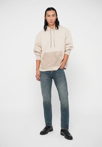 Bēša kapucejums ar teksturētu bēšu rakstu apakšā, apvienots ar zilām džinsām un melniem loaferiem. Gluda auduma un brīva piegriezuma.