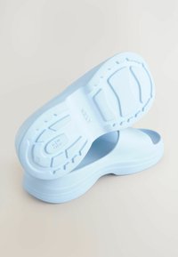 Ljusblåa slip-on skor med en texturerad gummisula. Slät, rundad design med öppen häl och minimala detaljer på sulan.