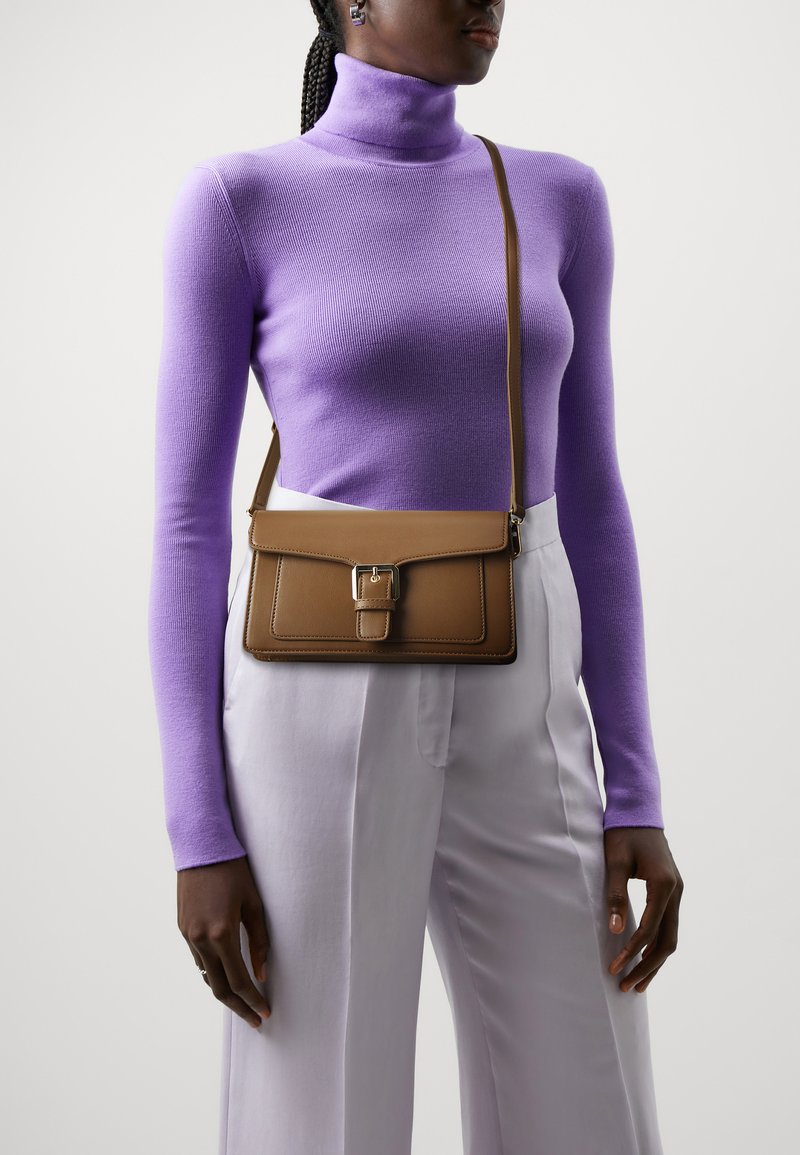 Brown leather crossbody bag with a rectangular shape, flap closure, and a metal clasp. Paired with a purple turtleneck and light-colored pants.