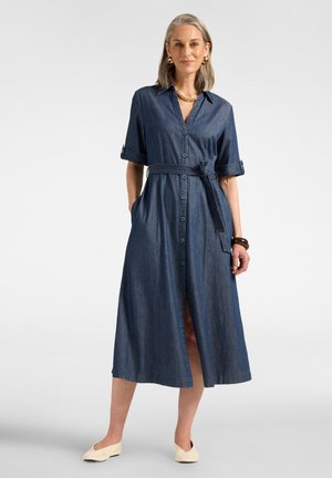 Donna in piedi che indossa un vestito midi blu con bottoni, maniche arrotolate, cintura annodata in vita, orecchini in oro, collana, braccialetto marrone e scarpe basse color crema.