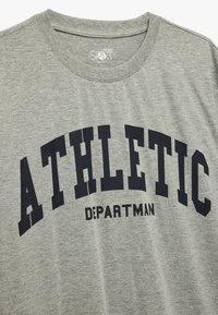 Szara koszulka z krótkim rękawem z nadrukiem, który głosi "ATHLETIC" dużymi czarnymi literami, z mniejszym tekstem "DEPARTMAN" poniżej.