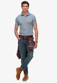 Jasnoniebieska koszulka polo, ciemnoniebieskie dżinsy, brązowe buty. Czerwona i czarna koszula w kratę związana w talii. Skórzany pasek i zegarek jako akcenty.