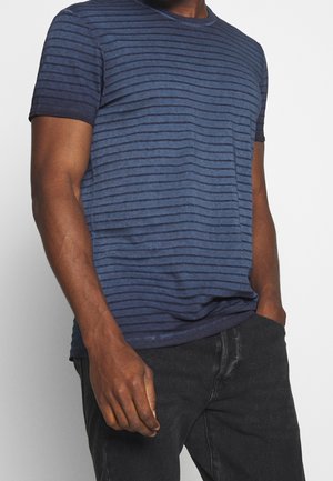 Man wearing a blue and black striped short-sleeve T-shirt with black jeans, one hand in pocket, against a plain white background.