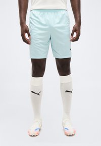 Άνδρας που φοράει ανοιχτό μπλε αθλητικά σορτς, λευκές κάλτσες Puma έως το γόνατο και λευκά ποδοσφαιρικά παπούτσια με μπλε και κόκκινες λεπτομέρειες, στέκεται μπροστά σε λευκό φόντο.