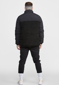 Man med nacktattoo iförd svart pufferjacka och fleecejacka, svarta byxor, vita strumpor och vita sneakers, står vänd bort från kameran mot en enhetlig bakgrund.