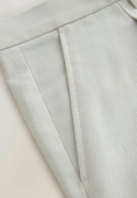 Gros plan d’un tissu gris clair texturé avec une couture verticale et un détail de poche fendue sur un vêtement.