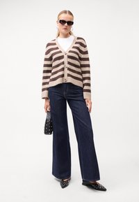 Καφέ και μπεζ ριγέ κα cardigan με κουμπιά, φορεμένο πάνω από λευκό τοπ. Συνδυασμένο με φαρδιά τζιν και μυτερές μαύρες γόβες, κρατώντας μια τσάντα με καρφιά.