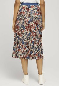Jupe plissée florale en crème avec des motifs rouges, bleus et noirs. Taille élastique, longueur genou, portée avec des bottines blanches.