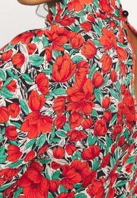 Nahaufnahme eines blühenden rot gemusterten Stoffes mit grünen Blättern auf einer Bluse mit hohem Kragen, die hinten mit einem schwarzen Knopf geschlossen wird.