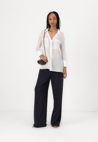 Camicia bianca trasparente con bottoni e due tasche sul petto, abbinata a pantaloni a gamba larga blu navy. Borsa a tracolla marrone e sandali neutri.