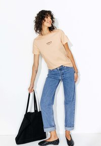 T-shirt en coton pêche avec imprimé "AMOUR", jean en denim bleu, sac fourre-tout en suède noir et ballerines noires à pois blancs. Cheveux bouclés coiffés.