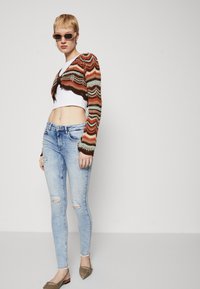 Knitted cropped cardigan in orange, brown, and green with wavy patterns over a white crop top; fitted light blue ripped jeans and flat shoes.