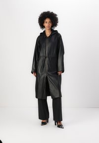 Palton lung din piele neagră, cu glugă, închidere cu fermoar pe față, talie elastică și buzunare laterale; asortat cu pantaloni corespunzători și tocuri negre.